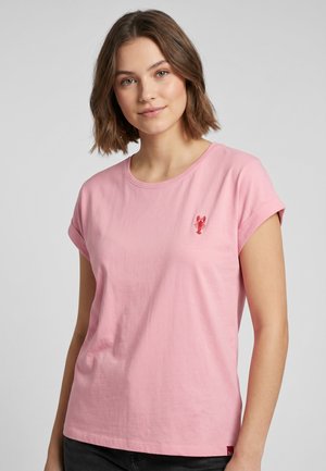 Junge Frau mit schulterlangem braunem Haar, die ein pinkes T-Shirt mit kleinem rotem Hummer-Emblem auf der linken Brust trägt.