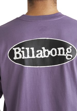 Camiseta de algodón morada con un gran logo ovalado negro y el texto blanco "Billabong" en la parte de atrás. Mangas cortas y ajuste relajado.