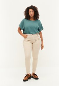 Turkusowa oversize bluzka z okrągłym dekoltem, w połączeniu z beżowymi, wysokim stanem dopasowanymi dżinsami oraz brązowymi butami sznurowanymi na grubej podeszwie.