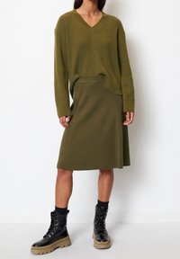 Olivgrüner, gerippter Pullover mit V-Ausschnitt, kombiniert mit einem passenden knielangen Rock. Schwarze Kampfstiefel mit dicken Sohlen runden den Look ab.