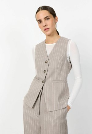 Mujer con cabello castaño recogido en una coleta baja, lleva un chaleco de rayas beige y pantalones a juego sobre una camiseta blanca de manga larga, de pie frente a un fondo blanco.