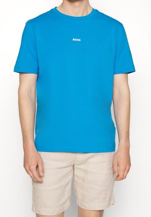 Mężczyzna w jasnoniebieskiej koszulce z krótkim rękawem z napisem „BOSS” na środku klatki piersiowej oraz beżowych szortach, stojący na białym tle.