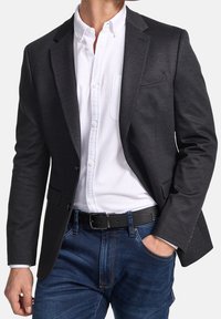 Tmavě šedý blazer z texturované látky, s úzkým střihem, klopovými límci a dvěma předními kapsami, nosí se přes bílou košili s knoflíky.
