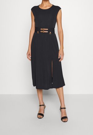 Femme portant une robe midi noire sans manches avec une fente à l'avant et une large ceinture à double boucle autour de la taille, associée à des talons noirs.