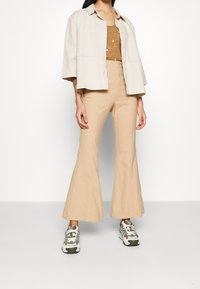 Beige kortärmad jacka över en brun topp, ställd med tanfärgade utsvängda byxor och sneakers med gröna accenter. Slät tyg med strukturerad design.