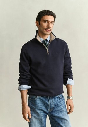 Man draagt een marineblauwe trui met een rits tot een kwart over een lichtblauw overhemd met opgerolde mouwen, een gestreepte stropdas, blauwe jeans en een zilveren polshorloge.