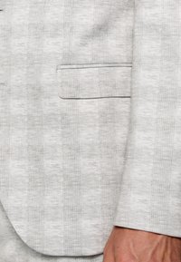 Sacou de costum cu carouri gri deschis, având un buzunar, textură subtilă și detalii fine, realizat dintr-un material ușor.