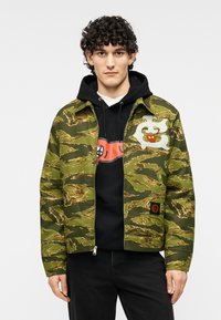 Ζακέτα καμουφλάζ με φερμουάρ σε πράσινους και καφέ τόνους, με υφή υφάσματος και κεντημένα γραφικά, φορεμένη πάνω από ένα μαύρο φούτερ με κουκούλα.