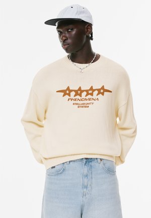Pulover crem cu grafică "STWD PHENOMENA STELLAR UNITY SYSTEM" în nuanțe de maroniu, asortat cu blugi albaștri deschisi și o șapcă gri. Material tricotat texturat.