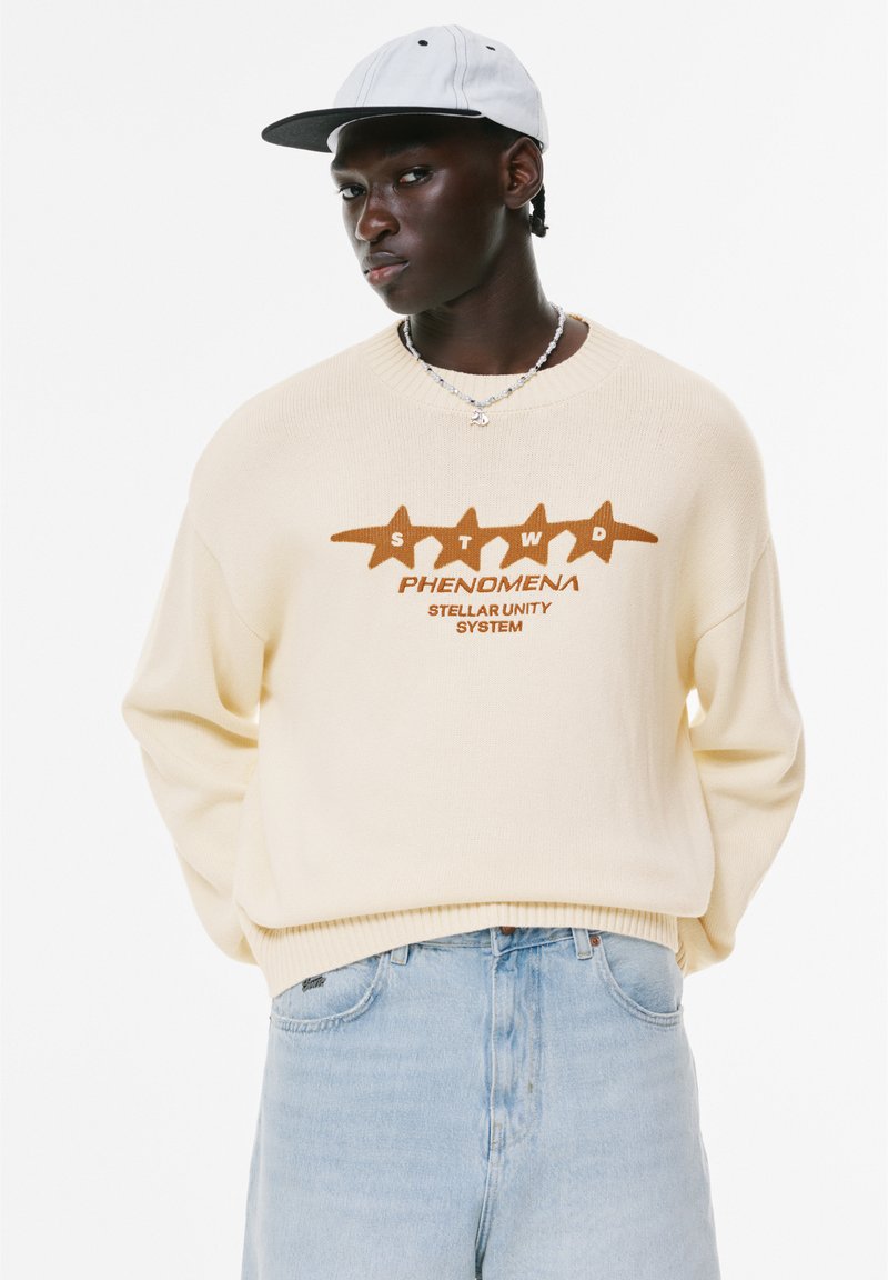 Pull crème avec un graphique "STWD PHENOMENA STELLAR UNITY SYSTEM" en brun, associé à un jean bleu clair et une casquette grise. Tissu tricoté texturé.