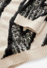Pintas vilnonių megztinis kreminės ir juodos spalvos. Jame yra raštuoti rankogaliai ir pūkuota tekstūra su abstrakčiu raštu.