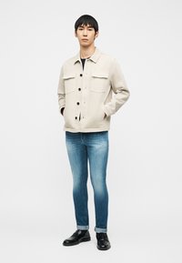 Man standing with hands in pockets wearing beige buttoned jacket, cuffed blue jeans, and black loafers against plain background.