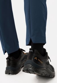 Black hiking shoes with orange accents, featuring a textured sole and mesh panels, paired with dark blue pants with a slit at the hem.