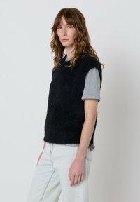 Moteris su banguotais rudais plaukais dėvi juodą pūkuotą be rankovių palaidinę virš pilkos trumpa rankovėmis marškinėlių ir šviesiai mėlynų džinsų, stovi prieš paprastą foną.