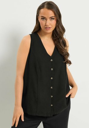 Vrouw met lang bruin haar, gekleed in een mouwloos zwart bloesje met knopen en een zwarte broek, poserend met één hand in de zak.