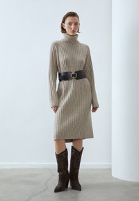 Abito in maglia a coste beige con collo alto, strizzato da una cintura in pelle nera con fibbia. Abbinato a stivali da cowboy ricamati marroni scuri.