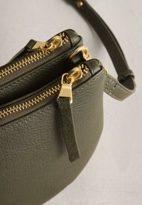 Sac bandoulière en cuir vert olive avec double fermeture éclair dorée et bracelet assorti sur fond neutre.