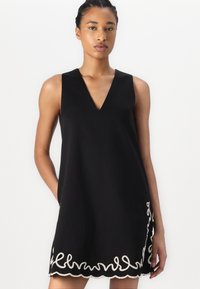 Black sleeveless dress with a V-neck, featuring a scalloped white embroidery design at the hem and a loose, A-line silhouette.