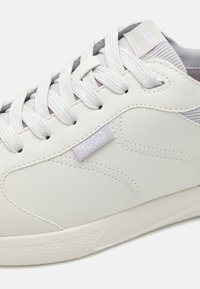 Witte leren sneaker met gestructureerde accenten, platte witte veters en een subtiele logo-tag. Heeft een gladde afwerking en een minimalistisch ontwerp.