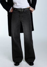 Personne portant un jean noir à jambes larges, une ceinture noire avec des œillets argentés, une chemise blanche, un manteau noir et des chaussures noires brillantes, debout les mains dans les poches.