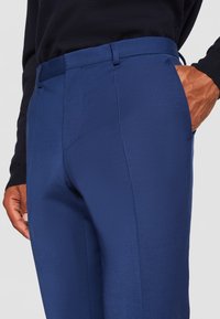 Blaue Stoffhosen mit glatter Textur, flachem Frontdesign, Seitentaschen und Gürtelschlaufen, kombiniert mit einem dunkelgrauen Pullover.