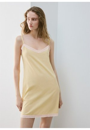 Ung kvinde med langt bølget hår iført en gul natkjole med lyserød blonderkant, stående foran en lys grå baggrund.