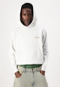 Grå hettegenser med frontlomme, med en liten gulllogo på venstre side. Båret over en grønn rutete skjorte, sammen med lyseblå jeans.