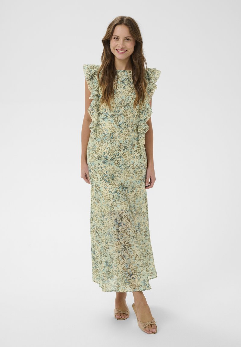 Mujer con un vestido largo sin mangas con estampado floral, hombros con volantes y zapatos de tacón abiertos beige, sonriendo y de pie sobre un fondo blanco.