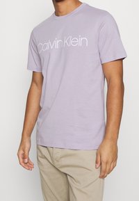 Camiseta de manga corta color lavanda claro, de algodón, con un logo blanco de "Calvin Klein" centrado en el pecho, diseño de cuello redondo.