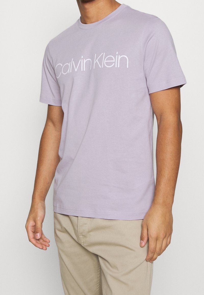 Camiseta de manga corta color lavanda claro, de algodón, con un logo blanco de "Calvin Klein" centrado en el pecho, diseño de cuello redondo.