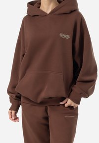 Sudadera marrón de tela suave con un corte holgado, bolsillo frontal y logo bordado. Puños y cinturilla acanalados para mayor textura.