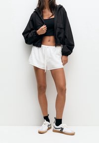 Veste noire courte avec capuche, associée à un short blanc texturé avec cordon de serrage. Stylé avec des baskets blanches et des chaussettes noires.