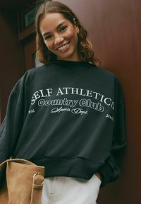 Felpa nera con testo bianco che recita "SELF ATHLETICS Country Club Sports Dept. est. 2023," dal fit rilassato e polsini a coste.
