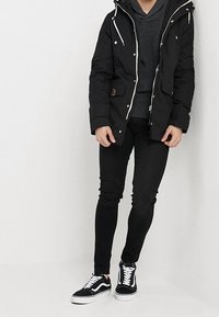 Černá parka s kapucí a bílými šňůrkami, ve spojení se šedým svetrem, černými úzkými džínami a černobílými teniskami.