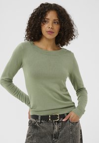 Ženska s kodrastimi lasmi, oblečena v svetlo zeleno dolgosleeve majico, zavedena v črne jeans hlače s kislinskim pranjem in črnim pasom, stoji ob beli ozadju.