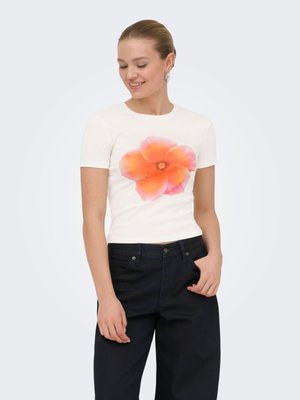 Mujer con camiseta blanca de manga corta con gran estampado de flores naranja y rosa, combinada con pantalones negros de talle alto, de pie contra fondo liso.