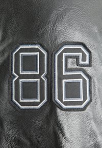Schwarzes Leder mit einer gestickten Nummer "86" in kontrastierenden weißen und blauen Fäden, auf einem strukturierten Hintergrund.
