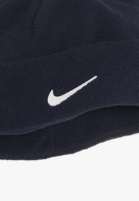Krnčasta modra flis kapa z belim Nike swoosh logotipom. Mehka tekstura z tesnim prileganjem, zasnovana za toploto in udobje.