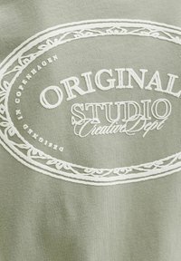 Heleda rohelise puuvillase tossusärk, millel on valge tikandiga logos tekst ja dekoratiivsed elemendid, pehme tekstuuri ja lõdva disainiga.