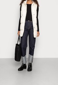 Langer weißer Puffer-Westen, schwarzer Rippstrickpullover, dunkle Jeans mit Denimabschlüssen, schwarze Tragetasche und schwarze Ankle-Boots.