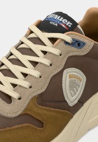 Sneaker marrone con sovrapposizioni in suede e pelle beige. Presenta lacci crema, un'etichetta con il logo e una linguetta imbottita con un accento blu.