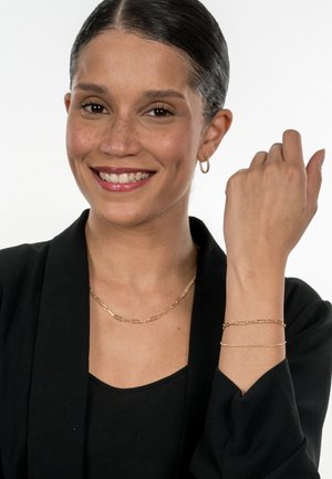 Goldschmuck an einem Model: eine Kettenhalskette mit länglichen Gliedern und ein dünner Drahtarmband, die beide eine schwarze Kleidung hervorheben.