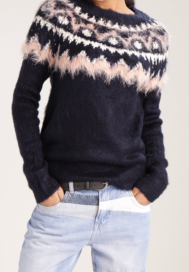 Marinblått fluffigt tröja med rosa, vit och svart geometrisk mönster över bröstet och axlarna, bärs med ljusblå lapptäckesjeans.