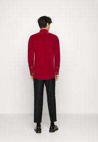 Czerwona koszula z długim rękawem, stojącym kołnierzem, mankietami na guziki i gładką fakturą, zestawiona z dopasowanymi czarnymi spodniami i błyszczącymi butami.