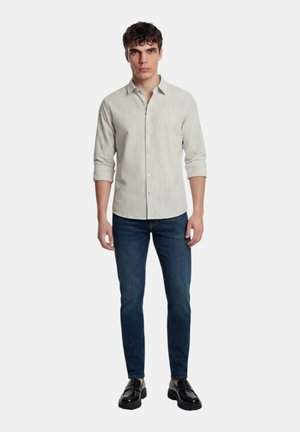 Giovane uomo che indossa una camicia leggera a righe con le maniche arrotolate, jeans blu scuro e mocassini neri, in piedi davanti a uno sfondo semplice.