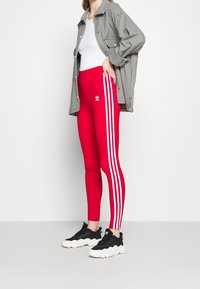Röda leggings med tre vita vertikala ränder, i kombination med en oversized svart och vit rutmönstrad skjorta samt svarta strukturerade sneakers.