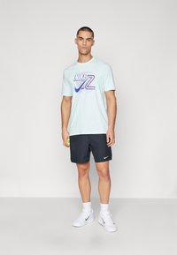 Camiseta de Nike de color azul claro con logo gráfico y pantalones cortos oscuros. Zapatillas blancas con detalles del logo en negro. Modelo sosteniendo una pelota de tenis verde.