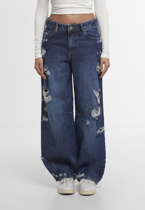 Loose dark blue distressed jeans with multiple rips worn with white sneakers and a cropped long-sleeve white top on a standing person.