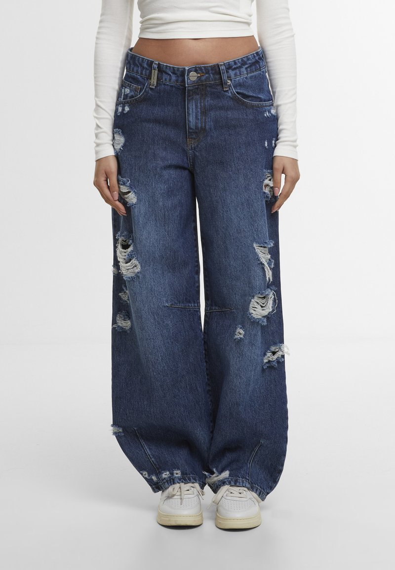 Jean bleu foncé ample et usé avec plusieurs déchirures, porté avec des baskets blanches et un haut blanc à manches longues court sur une personne debout.