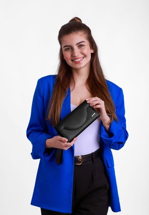Black textured handbag with a curved flap, three decorative dots, and an adjustable strap, held by a person in a blue blazer.